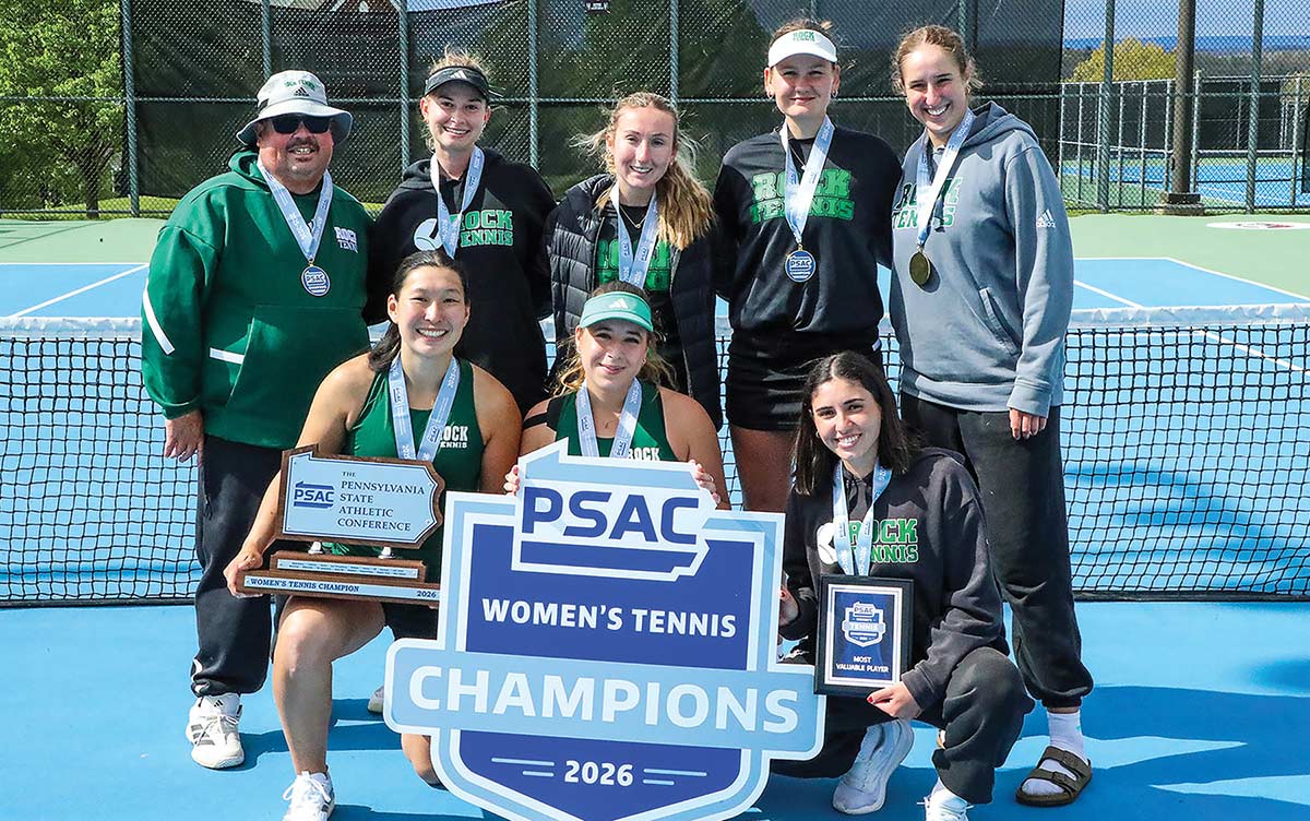 SRU women's tennis team individuals and their coach celebrating a championship victory in a group picture on an outdoor blue tennis court