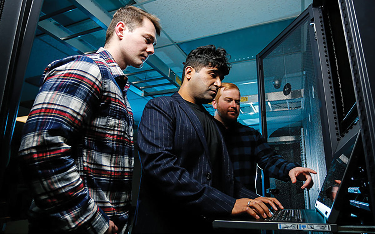 Three IT professionals working together inside a server room, with one man typing on a console laptop built into a server rack.