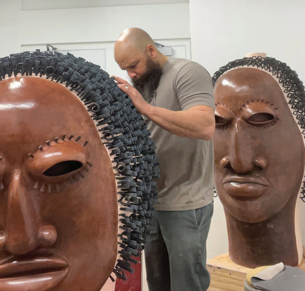 Sharif Bey works in his studio, adjusting the black textured hair on a large brown ceramic head sculpture. A second completed head sculpture stands in the background.
