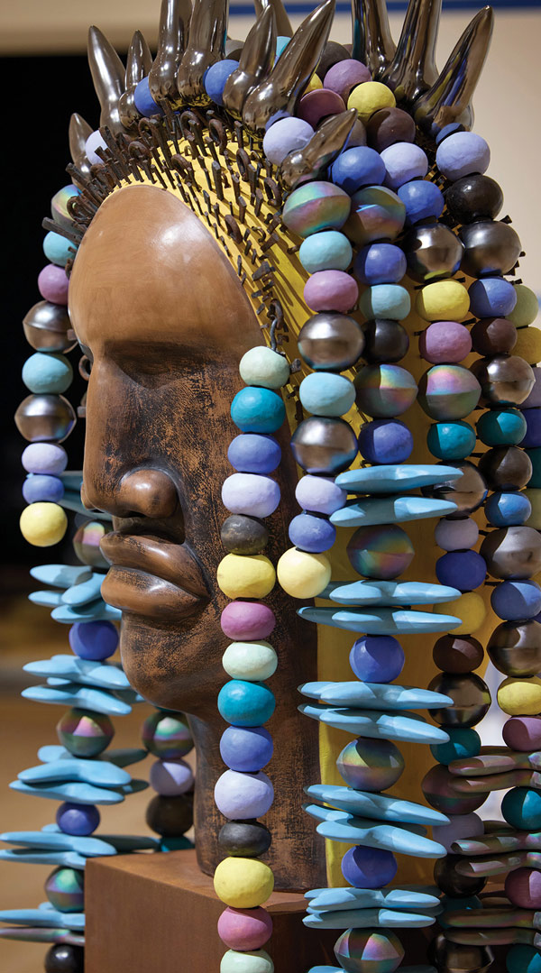 Profile view of Sharif Bey's sculpture "Crown Series: Iris," featuring a brown ceramic face, rows of colorful round and disk-shaped beads, and large dark spikes along the top.
