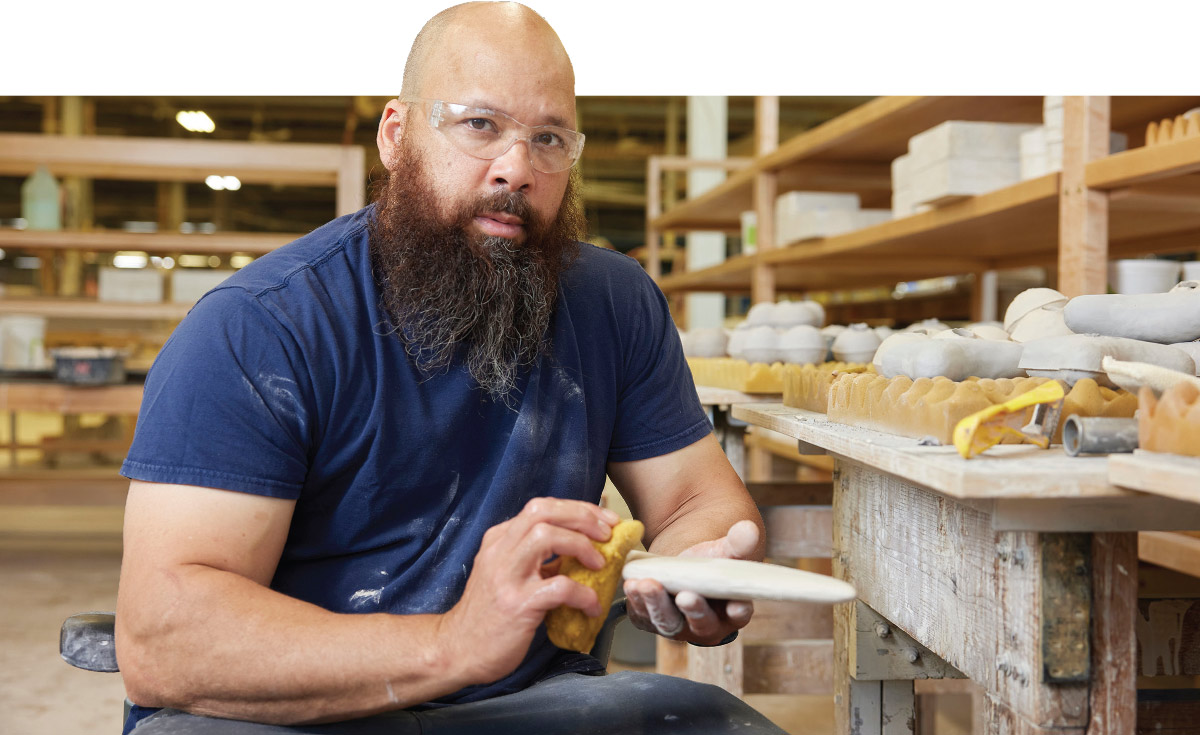 Sharif Bey, wearing safety glasses, sits in a studio holding a clay piece and a yellow sponge. Rows of white ceramic molds sit on wooden shelves in the background.
