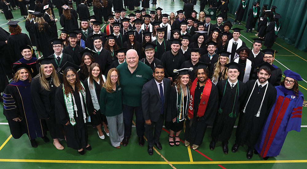 Elliott and Rhonda Haverlack, Prasad Vemala, faculty in regalia, and a large group of graduates in black caps and gowns stand together on a green gymnasium floor during a commencement ceremony.