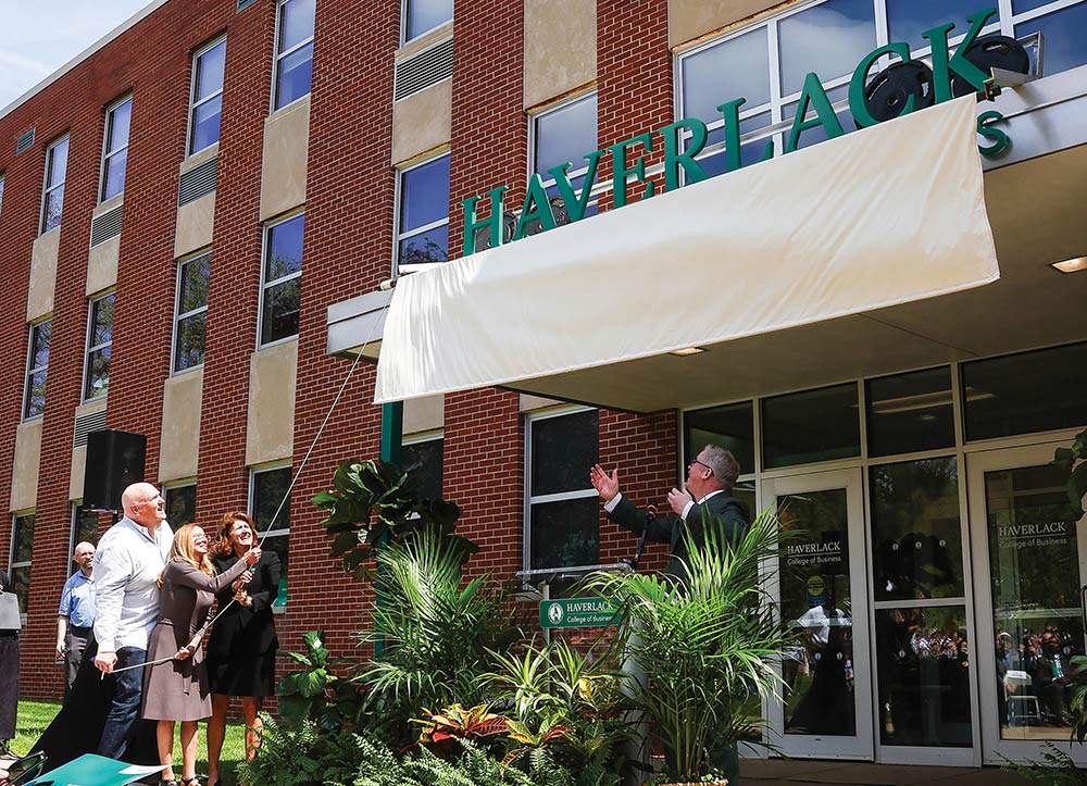 Elliott and Rhonda Haverlack, alongside SRU President Karen Riley, pull a rope to unveil the green "HAVERLACK" sign on a brick campus building while a man speaks at a podium surrounded by potted plants.