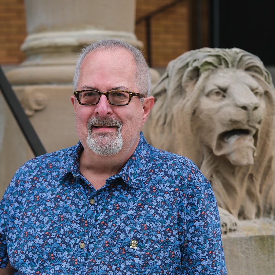Headshot of Ben Addison wearing a blue floral shirt and glasses, with a stone lion statue visible in the blurred background.