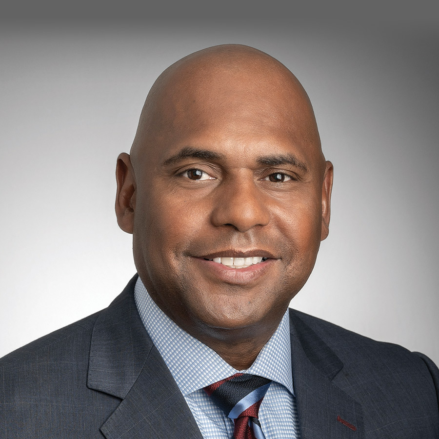 Professional studio headshot of Eric Conley smiling, wearing a gray suit, a light blue checkered dress shirt, and a striped tie against a gray background.