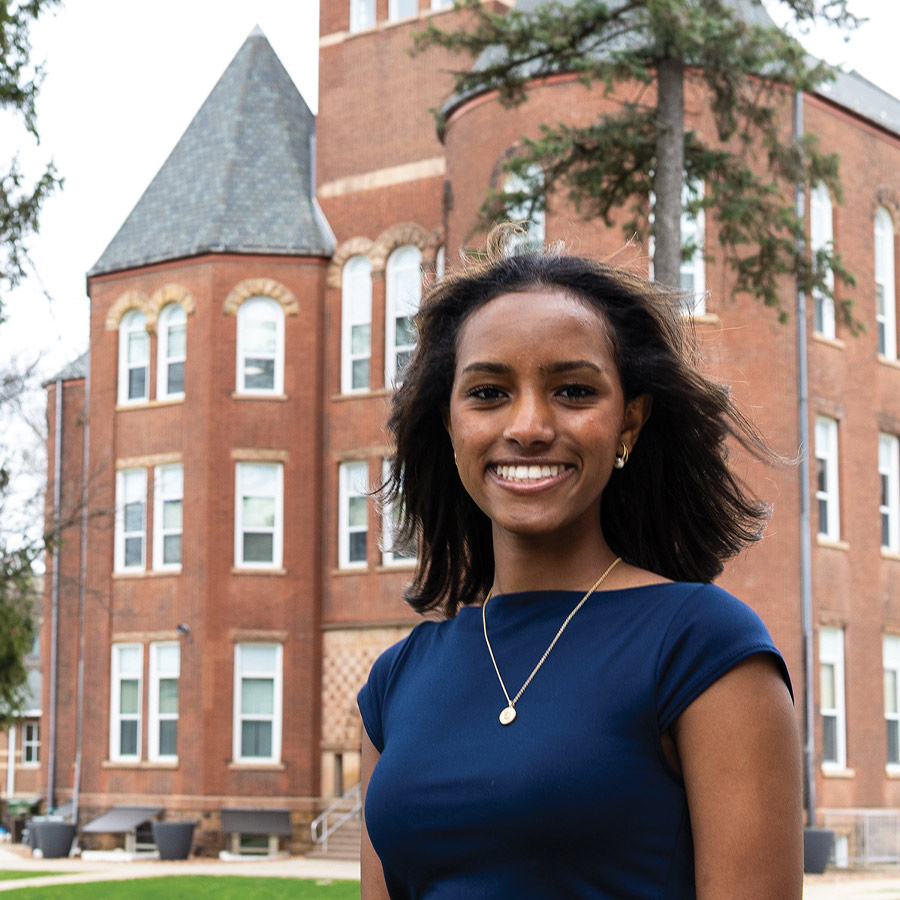 Headshot of Birtukan Hartman smiling and wearing a navy blue top and gold necklace, with a large red brick campus building in the background.