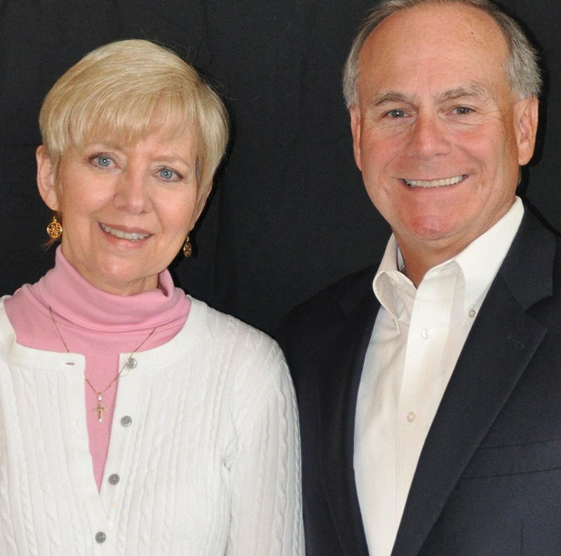 A smiling woman in a pink turtleneck and white cardigan poses closely with a smiling man in a navy suit jacket.