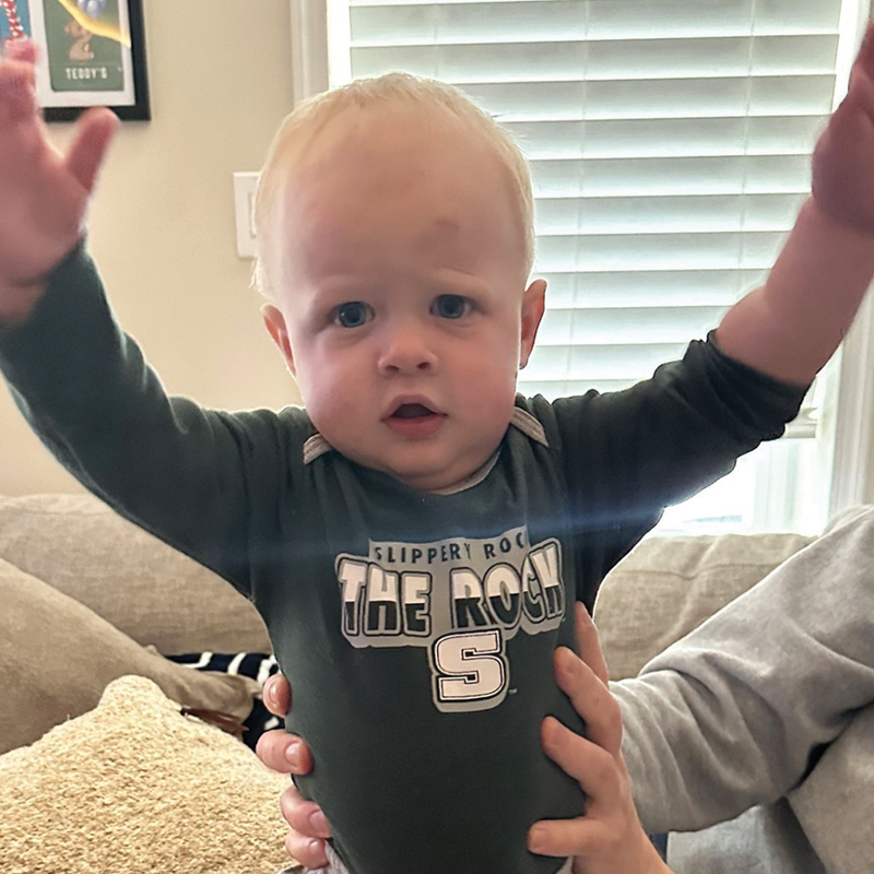 An infant wearing a green Slippery Rock "The Rock" onesie is held up playfully with arms raised in the air.
