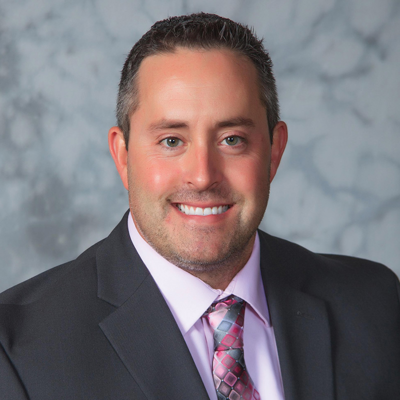 A formal studio headshot of a smiling man with short hair wearing a dark suit, pink shirt, and patterned tie.