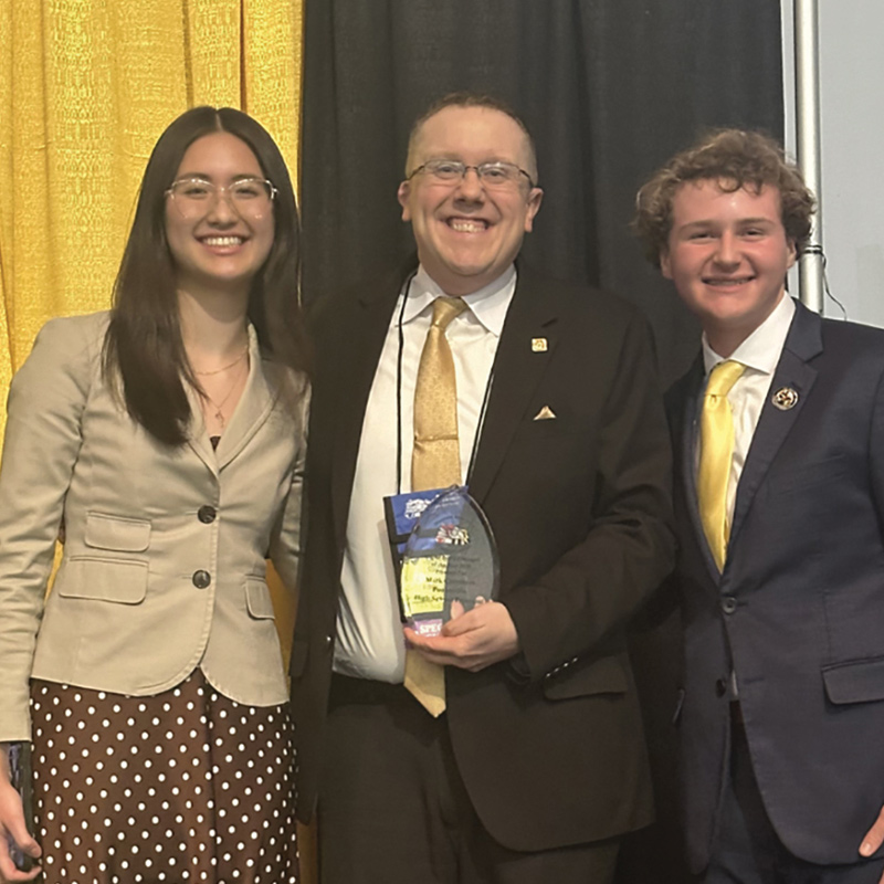 Three smiling individuals pose together indoors at an event while the man in the center holds a glass award.