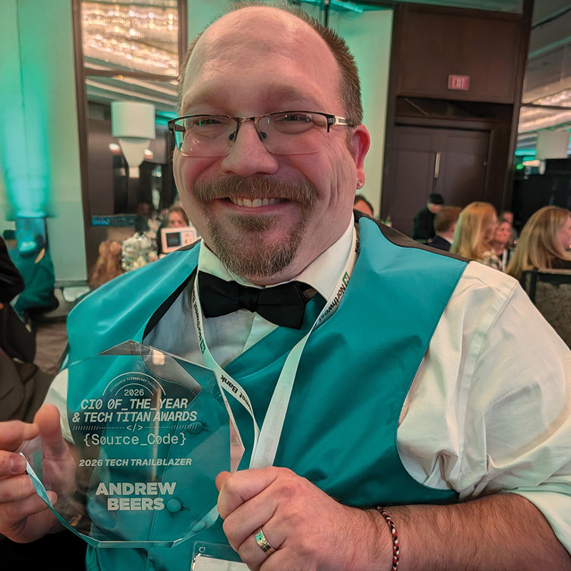A smiling man wearing glasses, a bow tie, and a teal vest holds up a glass "Tech Trailblazer" award plaque.