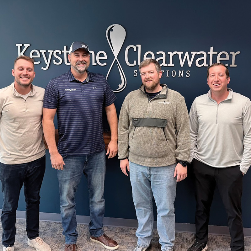 Four men stand in a row smiling in front of a blue wall featuring the "Keystone Clearwater Solutions" logo.