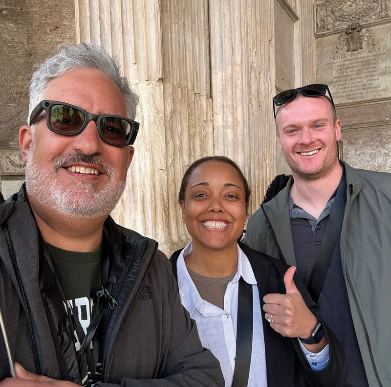 A selfie of three smiling individuals posing outside in front of a classic, light-colored stone column.