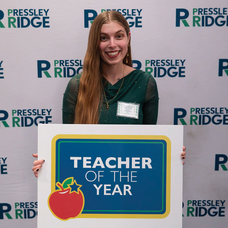 A smiling woman holding a large white sign that reads "Teacher of the Year" in front of a branded backdrop.