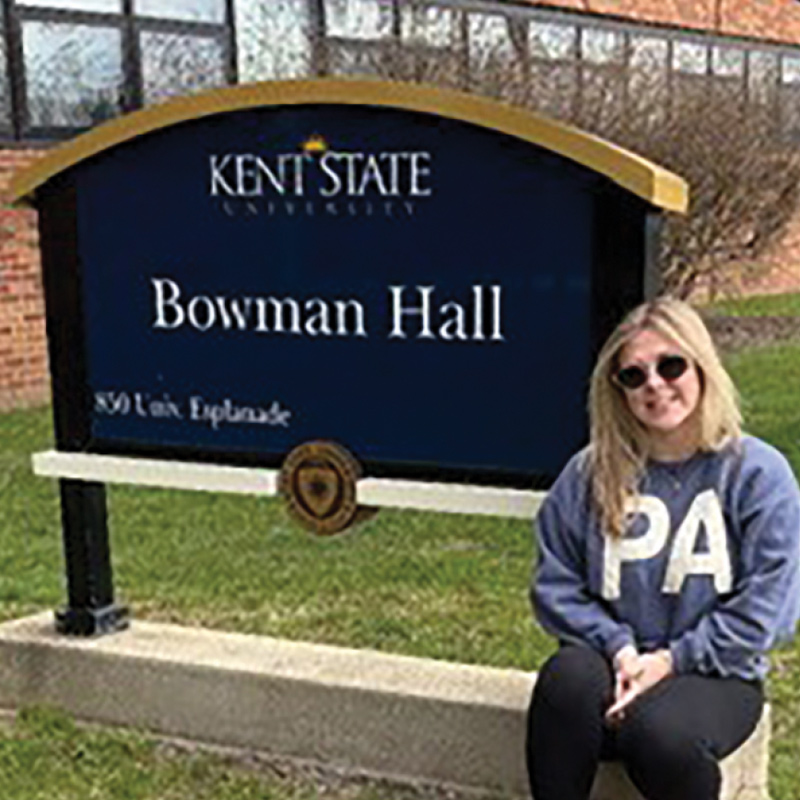 A woman wearing sunglasses and a grey sweatshirt sits outside next to a Kent State University Bowman Hall sign.