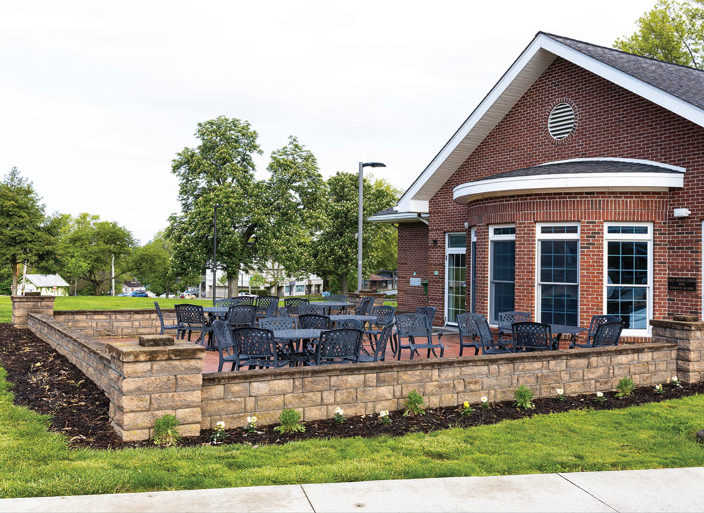 An outdoor brick patio area next to a building filled with dark metal tables and chairs behind a low stone wall.