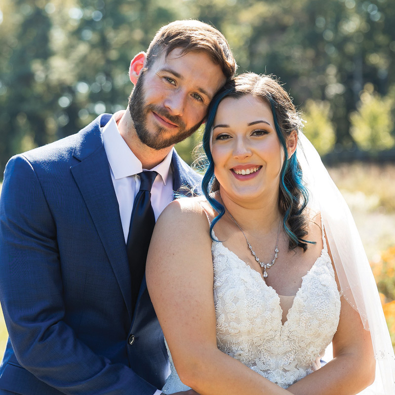 A groom in a blue suit rests his head against his smiling bride, who wears a lace wedding gown and a veil.