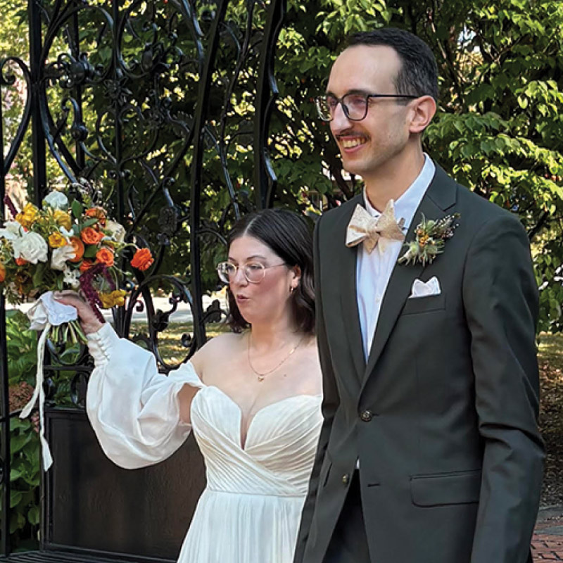 A bride in an off-the-shoulder gown holds up a vibrant bouquet next to a groom in a dark suit and bow tie.