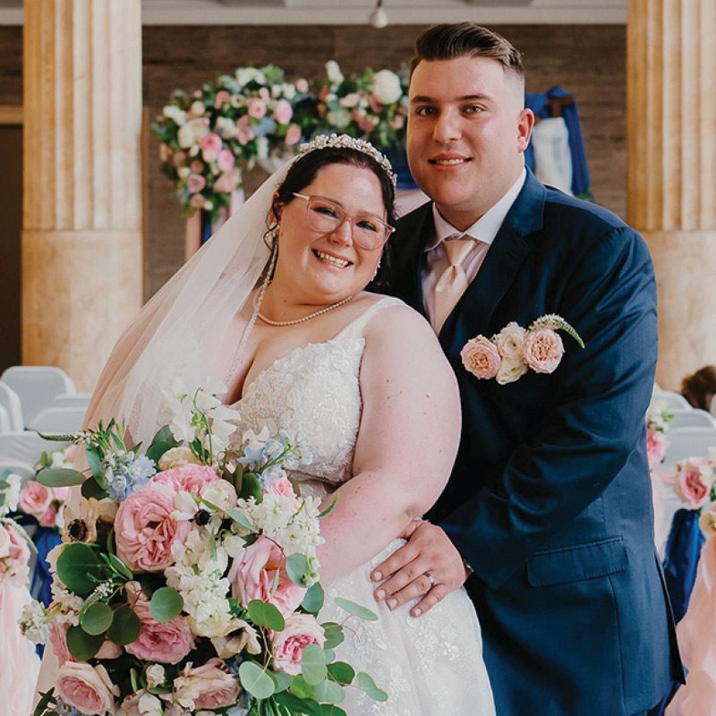 A bride wearing a tiara and glasses smiles warmly as her groom in a navy suit embraces her from behind.