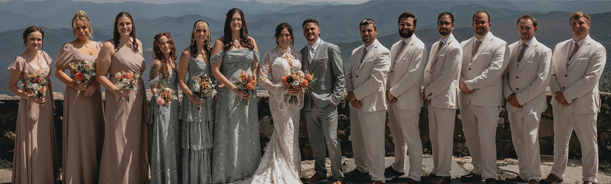 A full wedding party poses together outdoors in a row against a scenic background of misty rolling mountains.
