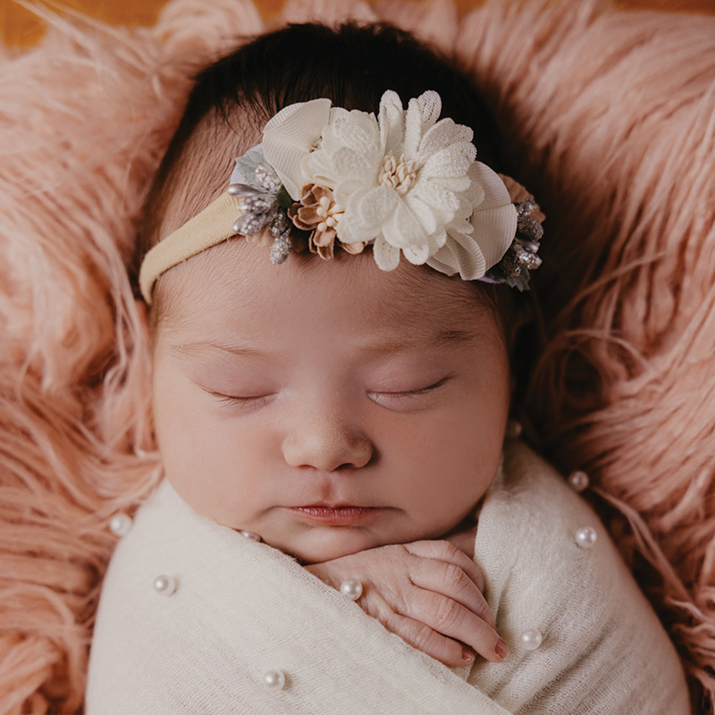A sleeping newborn baby swaddled in a white pearl-accented blanket wears a large white floral headband.