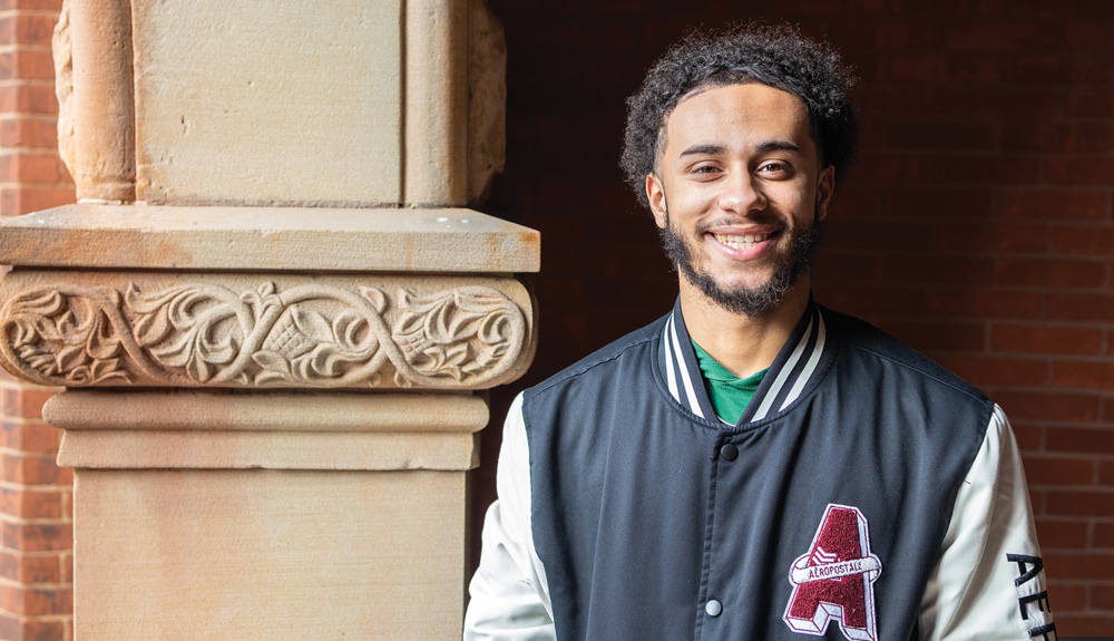 A smiling young man with curly hair wearing a varsity jacket poses next to a carved stone pillar.