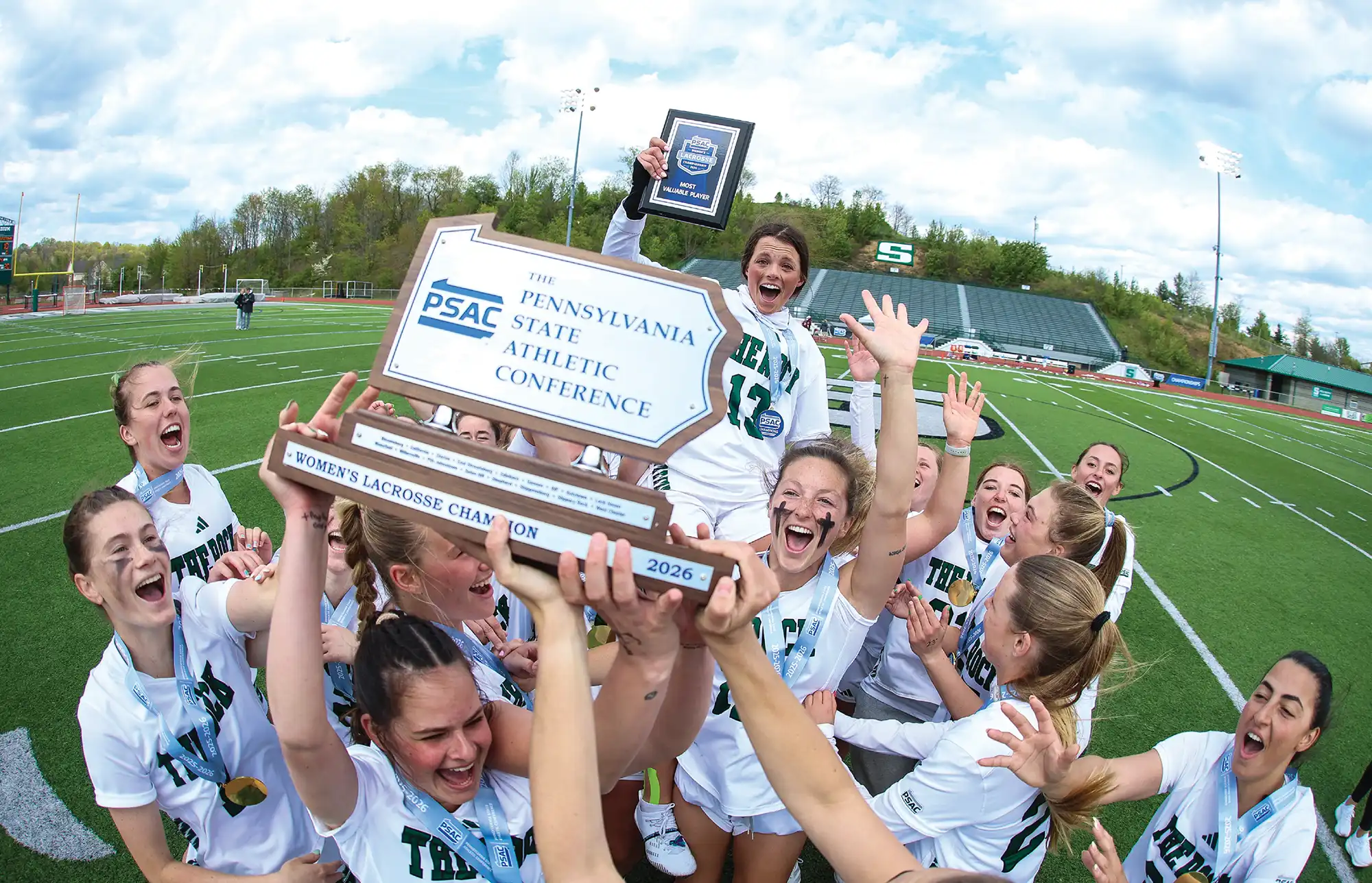 The Slippery Rock women’s lacrosse team ecstatically celebrates on the field while hoisting their 2026 PSAC championship trophy.