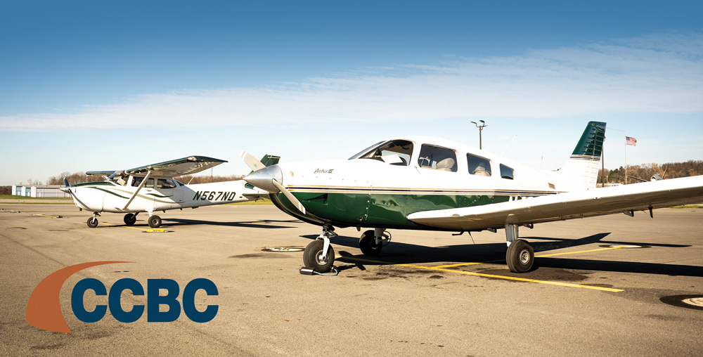 Two small propeller airplanes parked on an airport tarmac under a clear sky. The airplane in the foreground is white and dark green, while a second white and green plane is visible in the background.