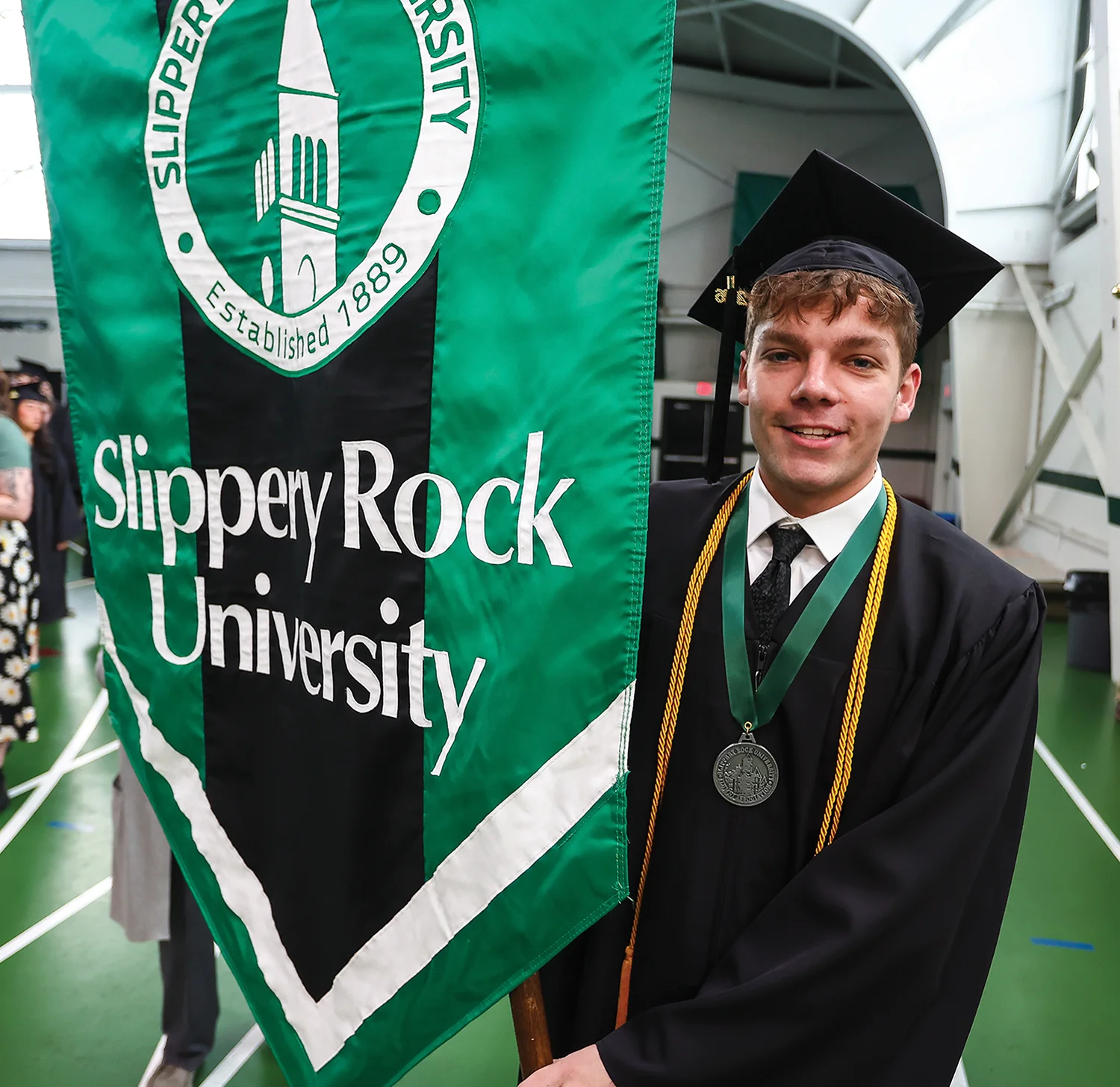 A male graduate in a cap and gown smiles while holding a large green Slippery Rock University ceremonial banner.