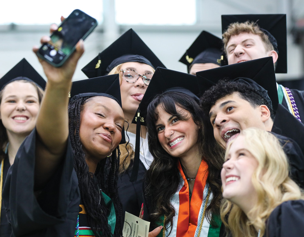 A joyful group of Slippery Rock graduates in caps and gowns crowd together to lean in and smile for a selfie.