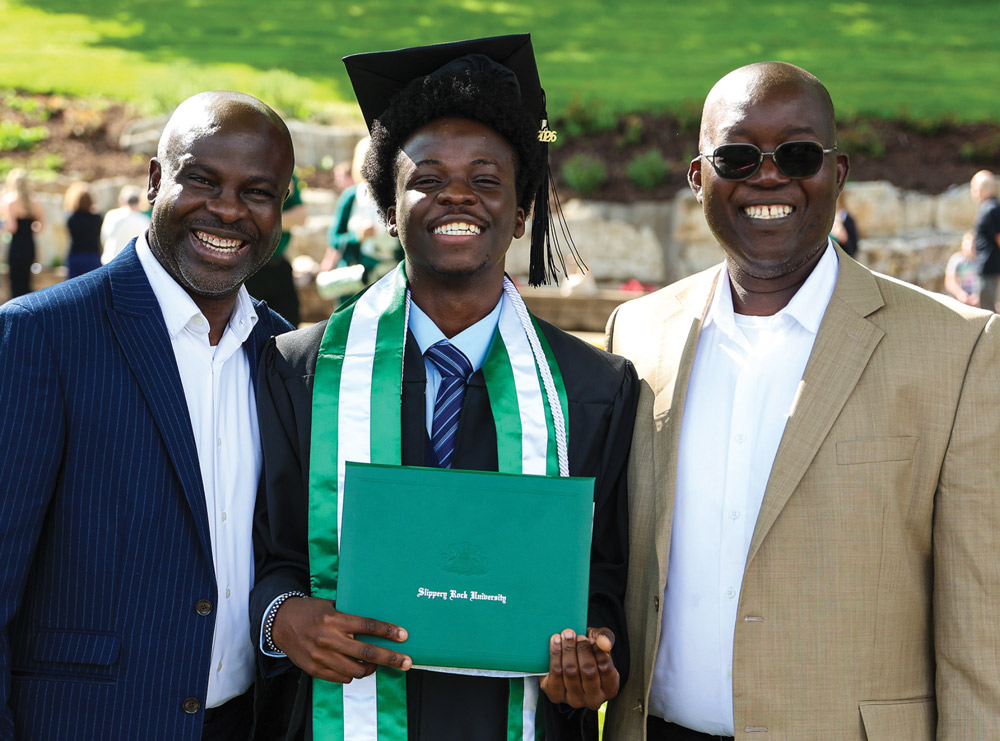A happy male graduate holds his green Slippery Rock diploma while posing outside between two smiling older men.