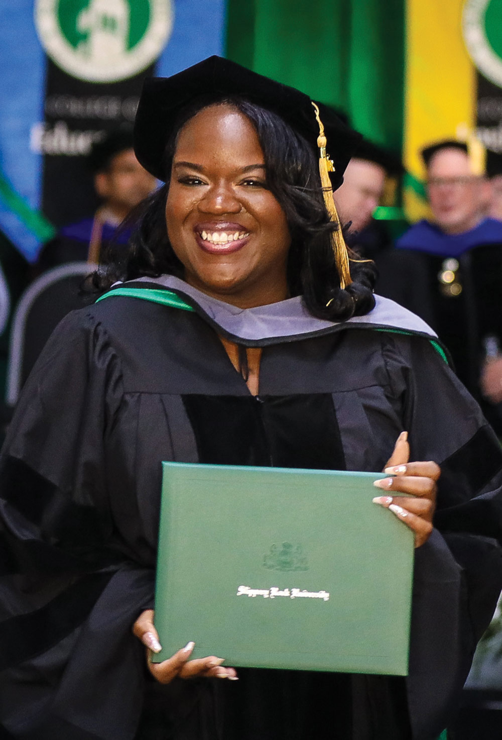 A smiling female graduate wearing doctoral regalia proudly holds up a green Slippery Rock University diploma.