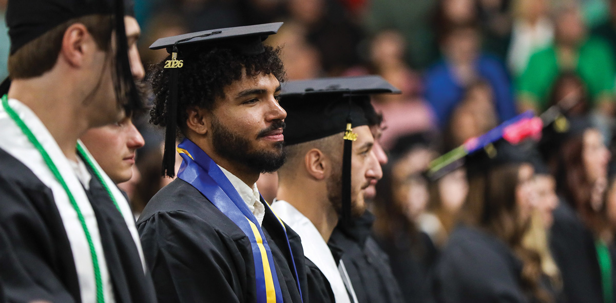 A row of Slippery Rock graduates in caps and gowns sit in profile view during a commencement ceremony.