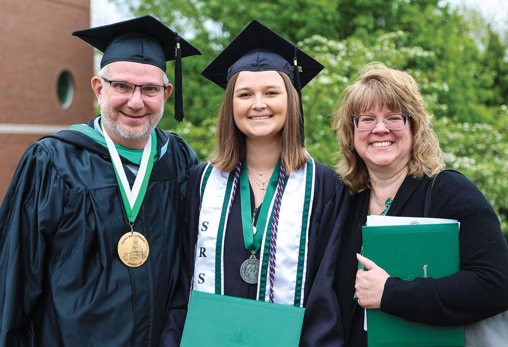 A female graduate poses outside with two proud family members while holding her green Slippery Rock diploma.