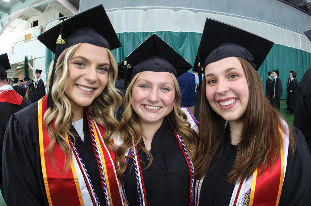 Three smiling female graduates in black caps and colorful stoles pose closely together at a ceremony.