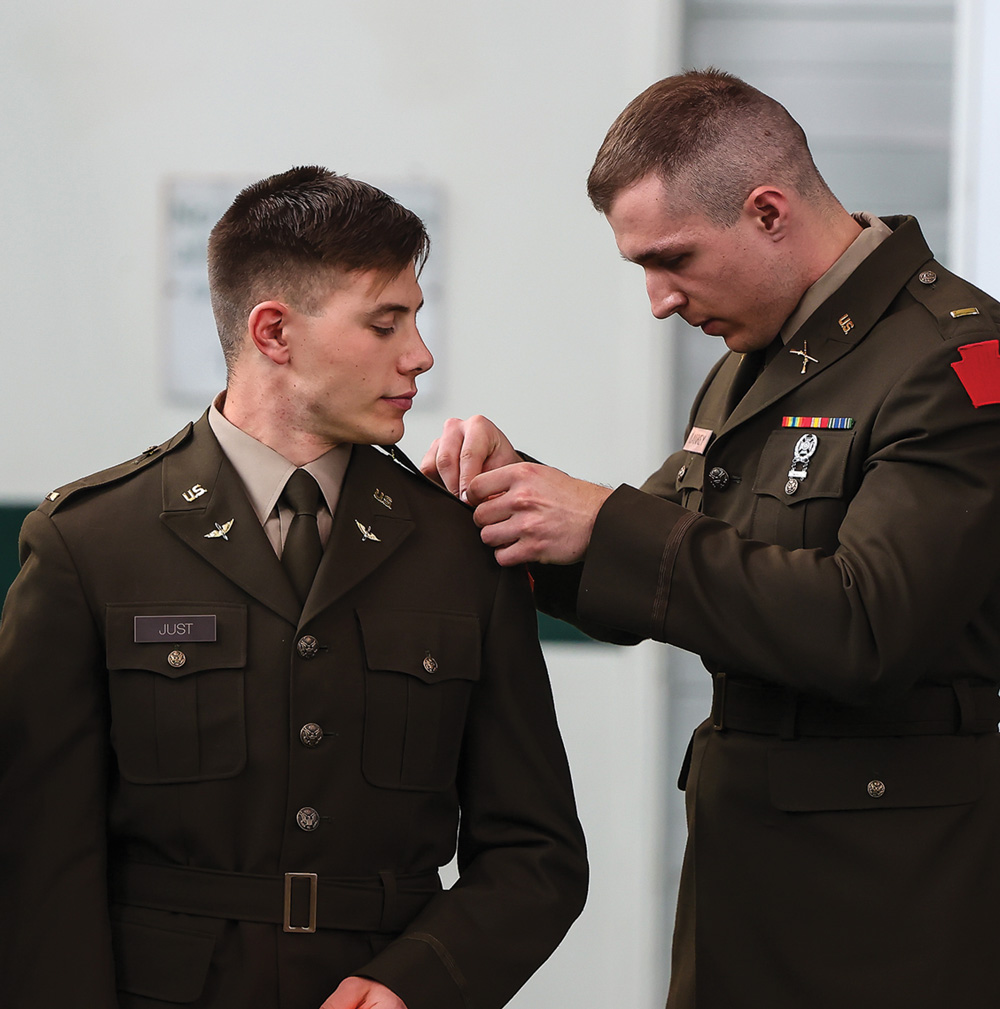 An army ROTC service member adjusts the shoulder insignia on another soldier’s dress uniform.