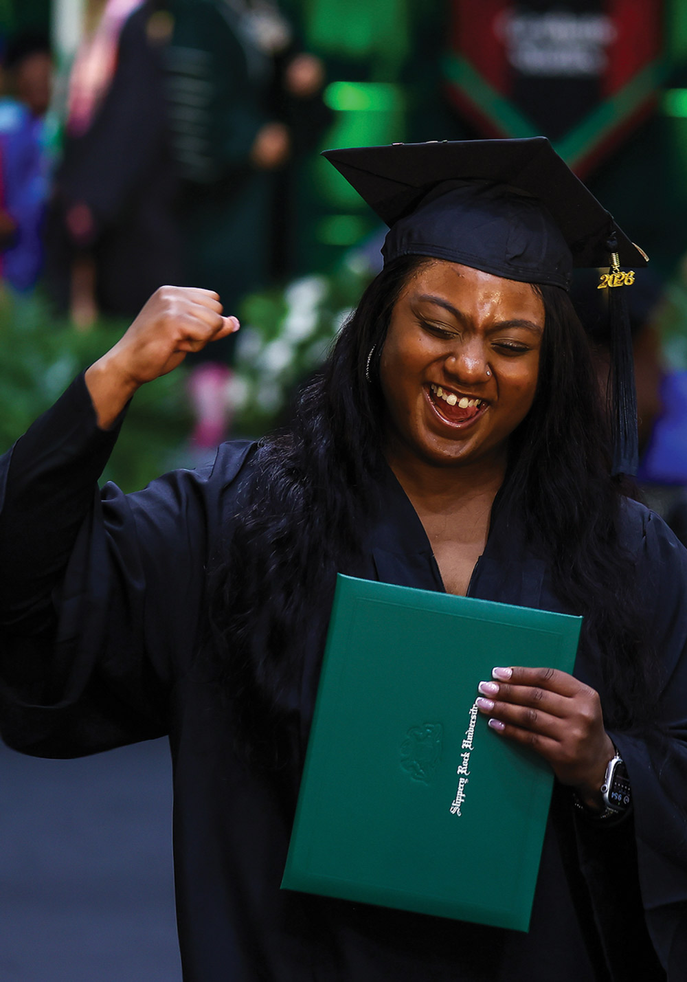 An excited female graduate cheers and raises her fist while holding her green Slippery Rock diploma.