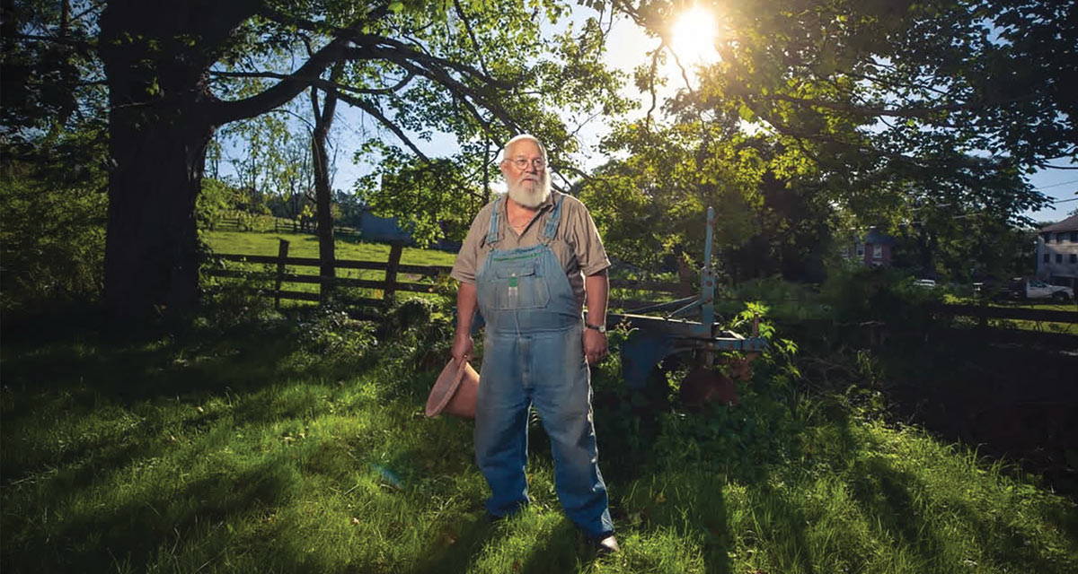 An older man with a long white beard wearing denim overalls stands in a grassy field beneath large shade trees with bright sunlight.