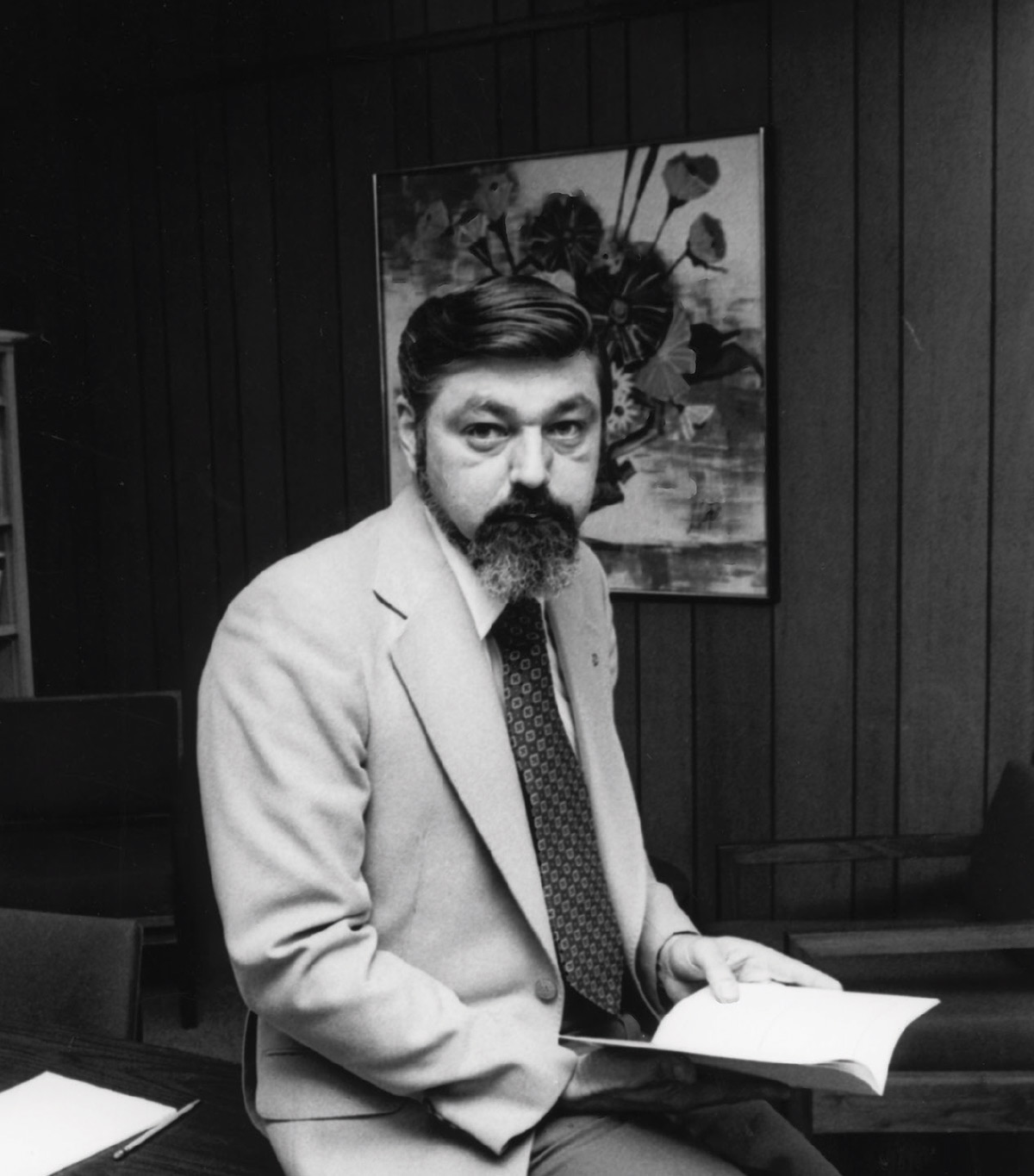 A black-and-white portrait of a man with a beard and mustache wearing a light suit jacket and tie while holding an open book.