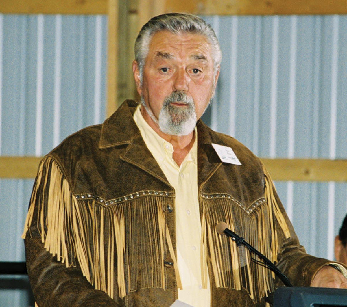 A man with a grey beard and mustache speaking at a podium while wearing a brown suede jacket featuring western-style fringe.