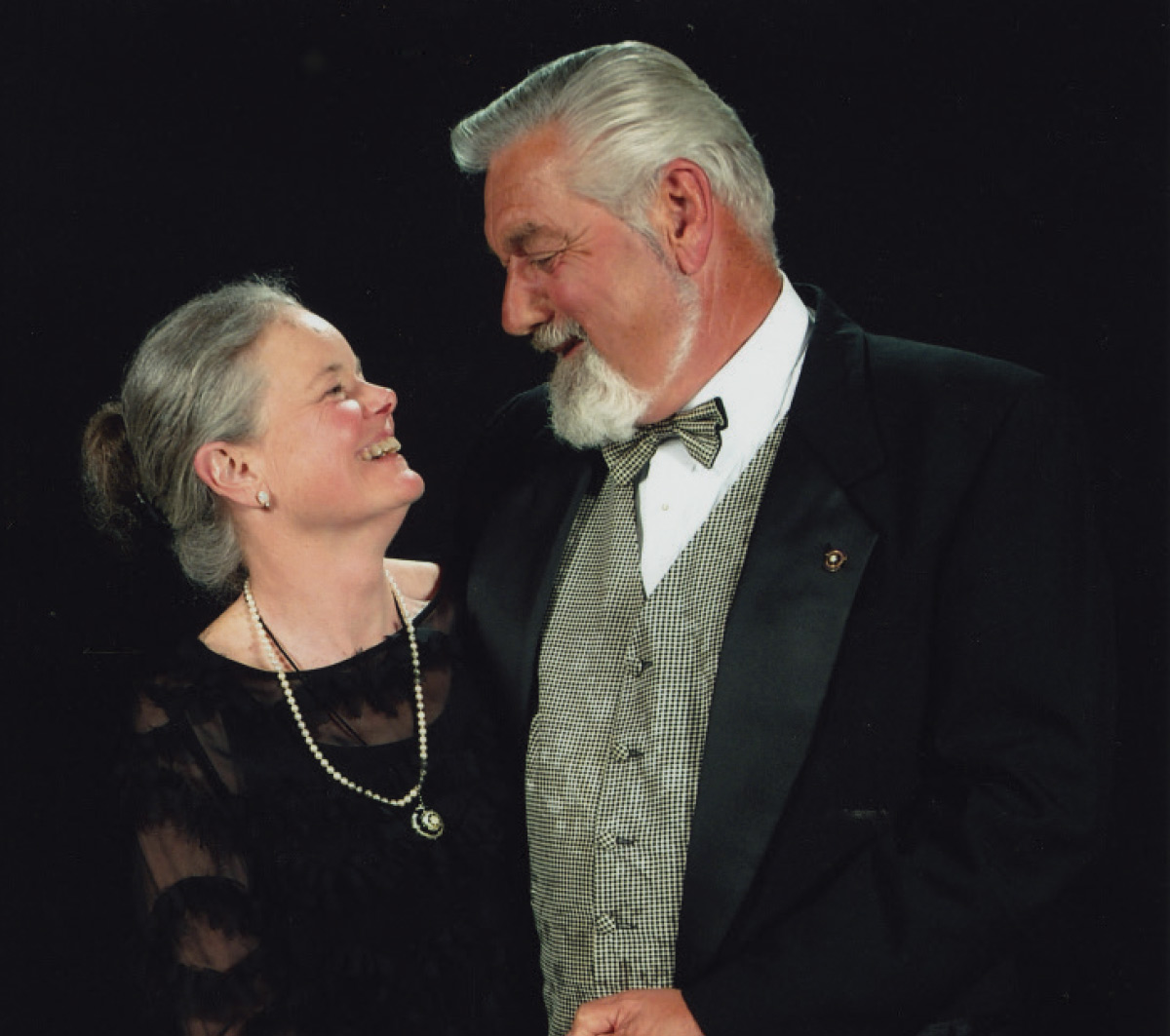 A smiling older couple looking affectionately at one another against a dark background, with the man in a tuxedo and bow tie.