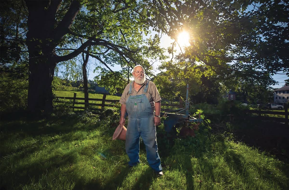 An older man with a long white beard wearing denim overalls stands in a grassy field beneath large shade trees with bright sunlight.