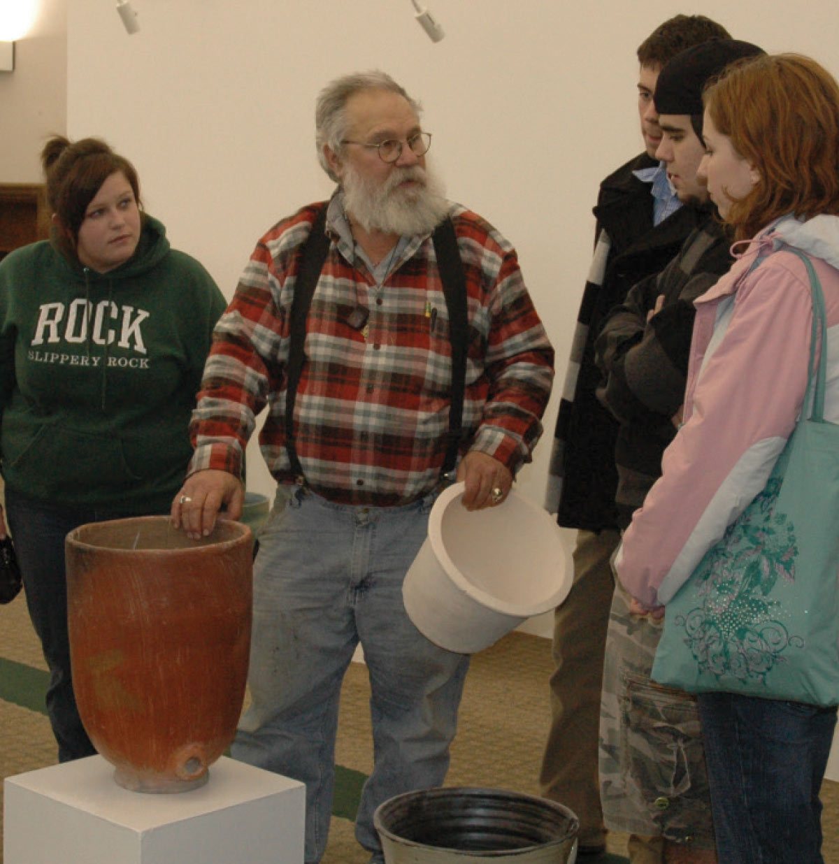 An older bearded instructor in a flannel shirt and suspenders demonstrates pottery techniques to a group of university students.