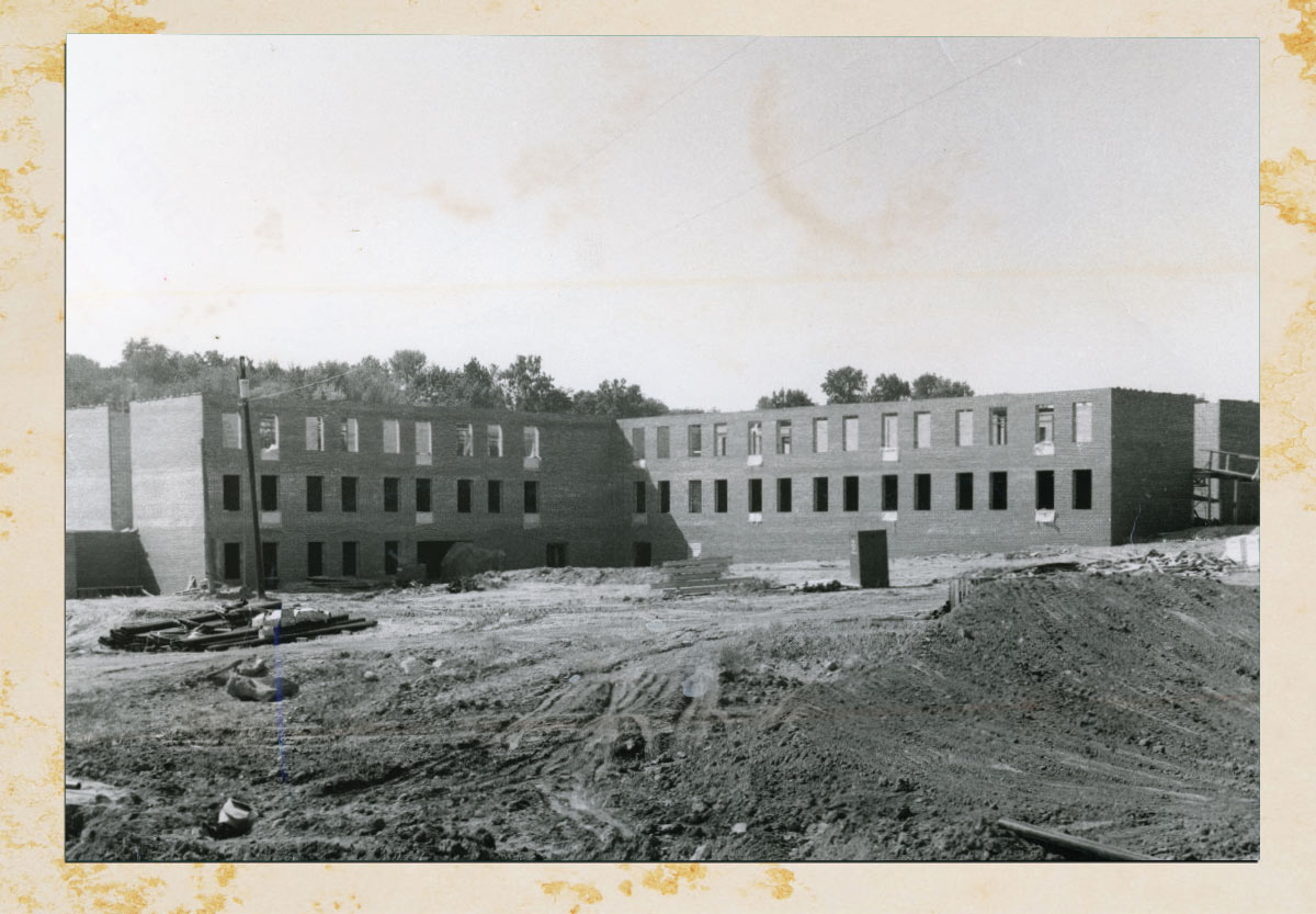 A vintage black-and-white photograph capturing the structural brick shell of a large institutional building under construction on a dirt lot.