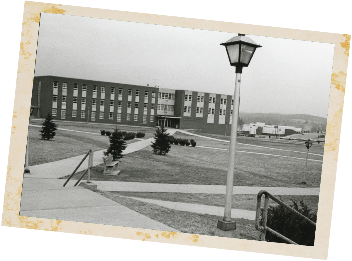 A wide, angled black-and-white photo of a university building and its sprawling lawn, framed by a prominent streetlamp in the foreground.