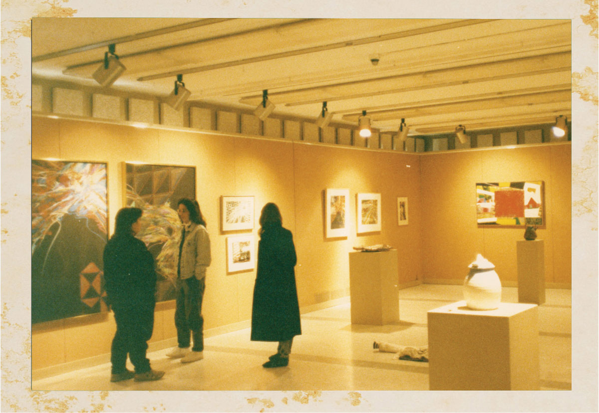An indoor shot with warm lighting showing visitors viewing paintings and sculptures displayed on pedestals inside an art gallery.