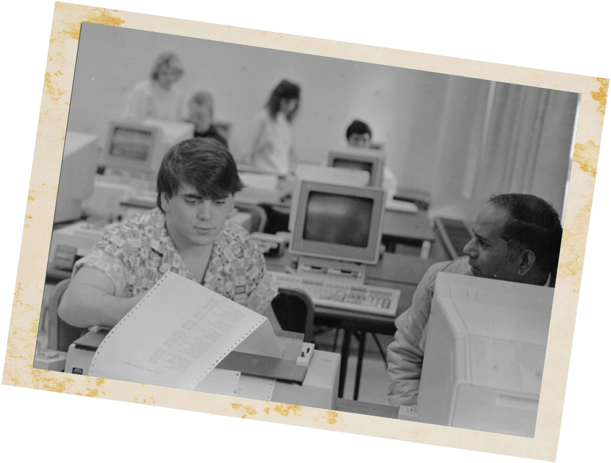 A black-and-white photo from a computer lab showing an instructor assisting a student who is looking over a continuous feed paper printout.