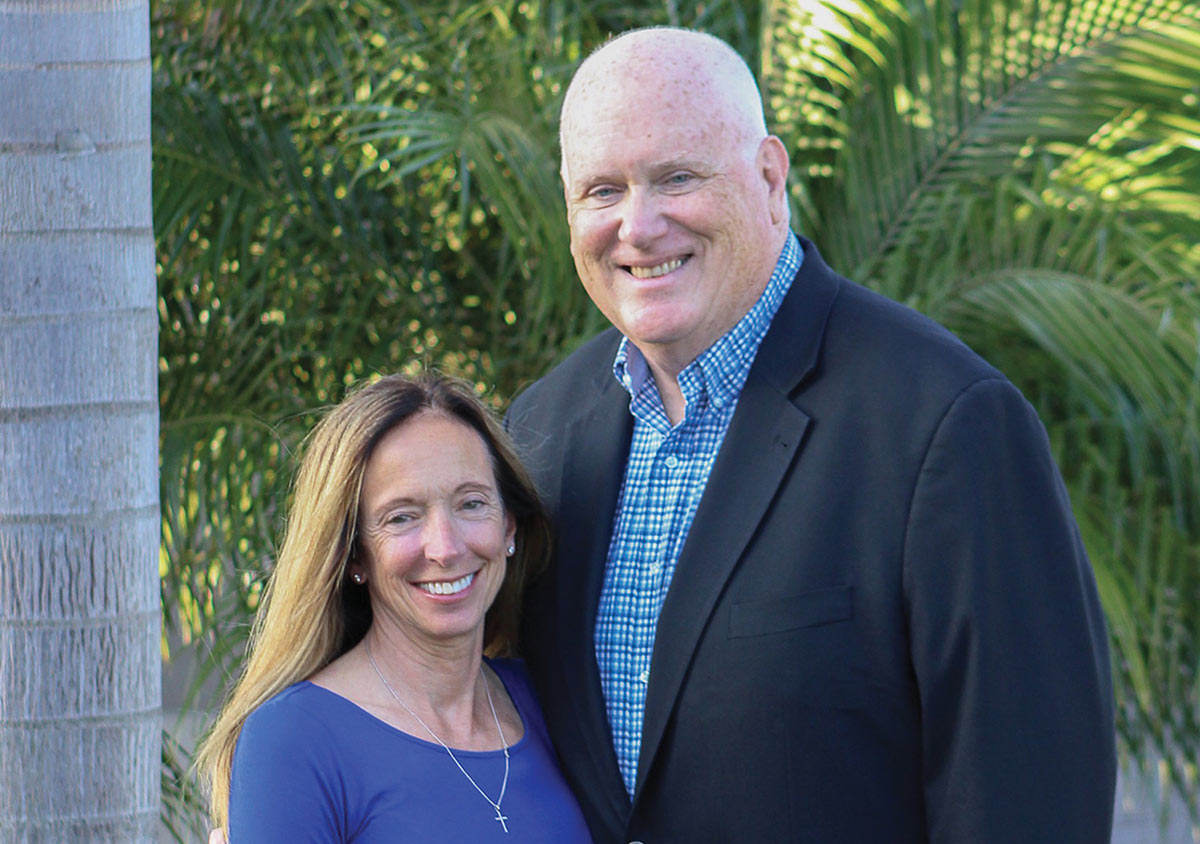 A smiling man in a dark blazer and a woman with long hair in a blue top pose together outdoors in front of lush green palm foliage.