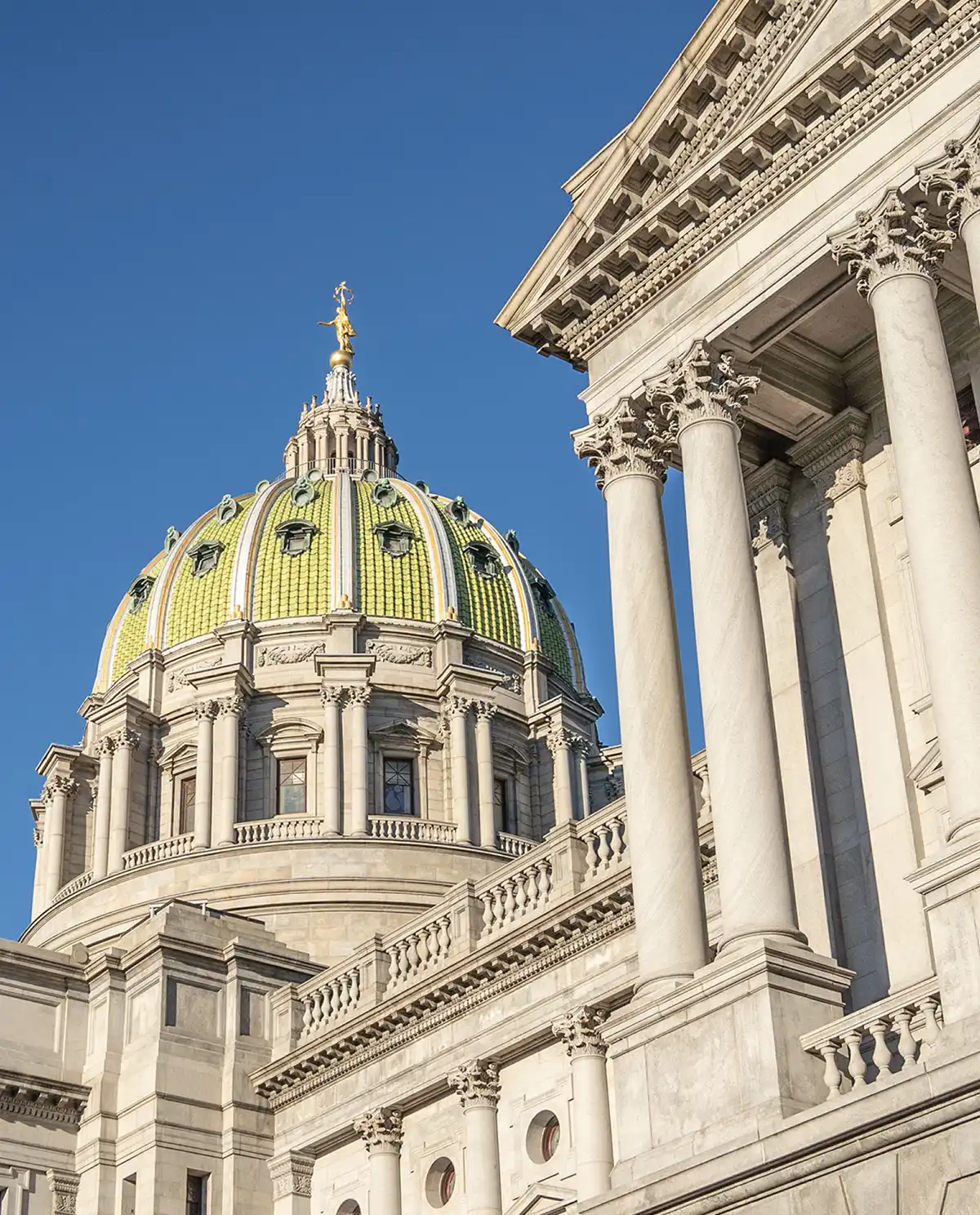 The ornate dome of a capitol building features green tiling and a gold statue at its peak, viewed past large classical stone columns under a clear blue sky.