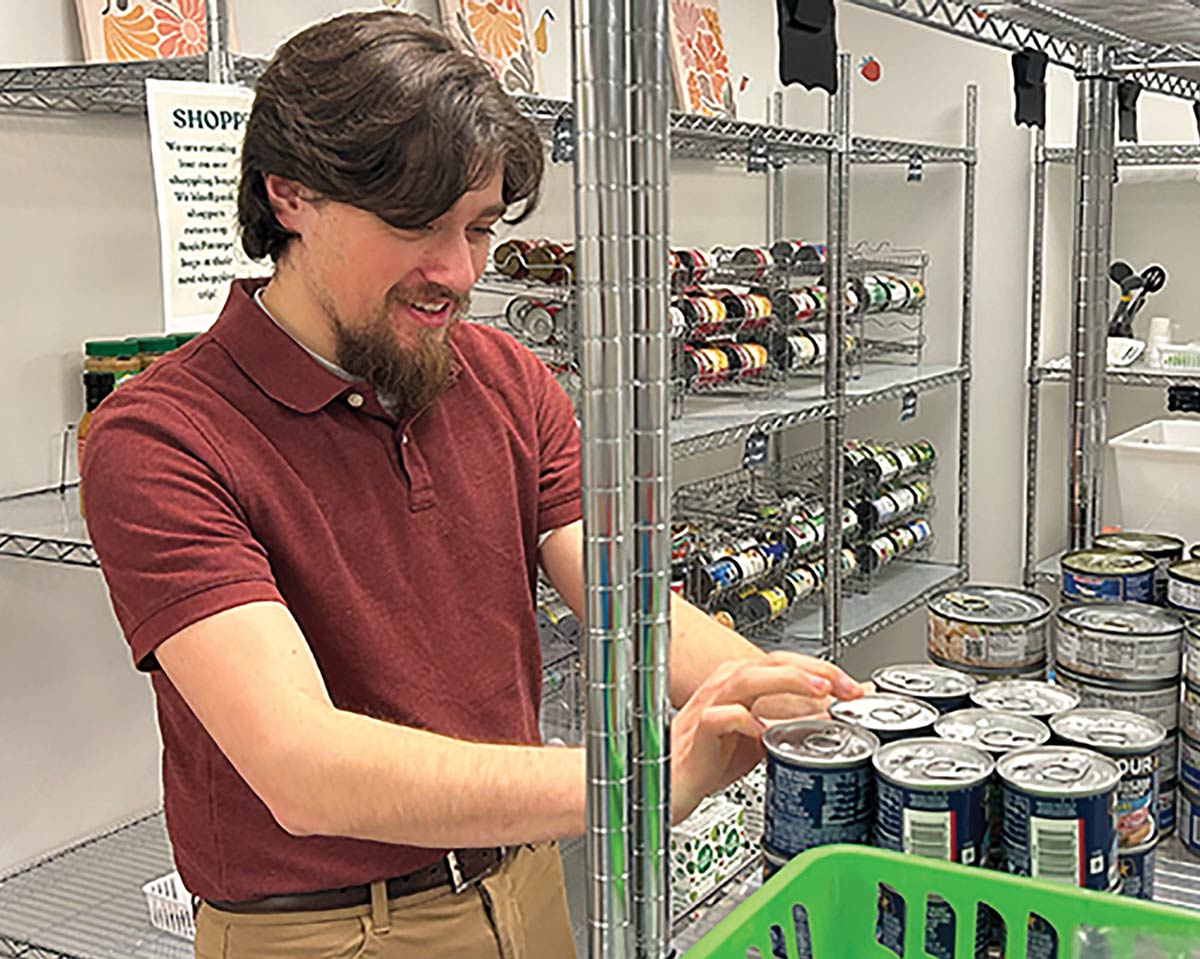 A smiling man in a maroon polo shirt organizes stacks of canned goods in a food pantry filled with metal wire shelving.